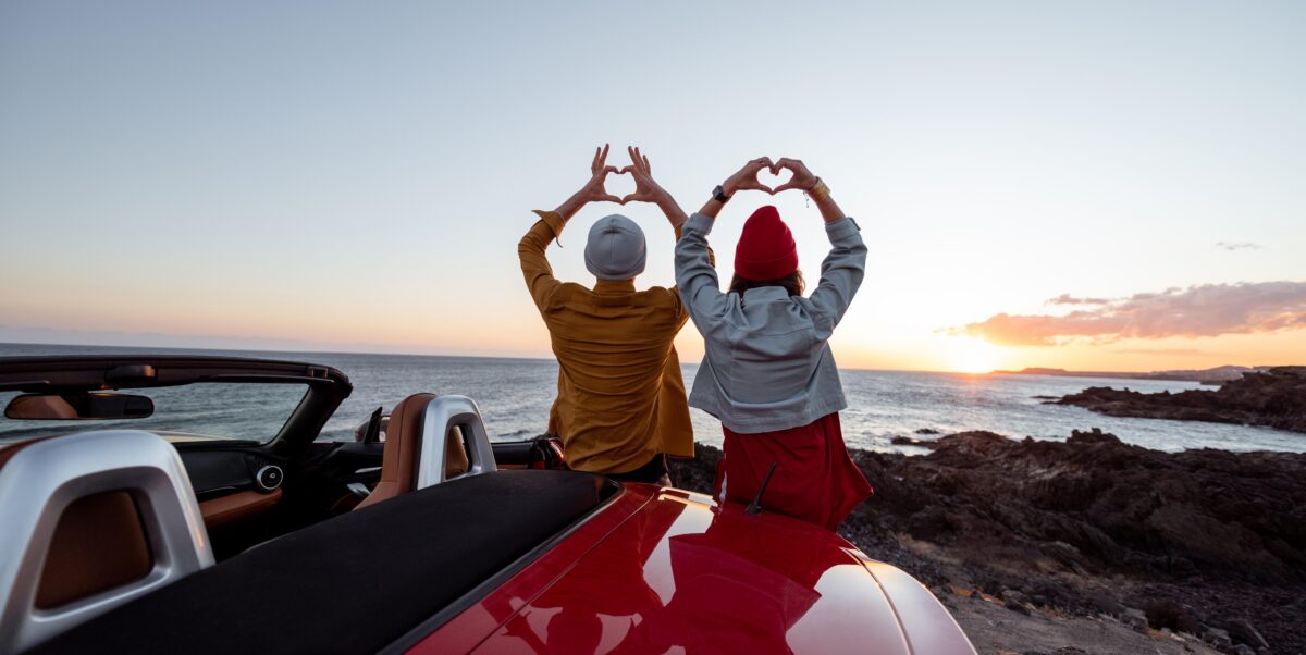 Two people leaning against a car holding up their hands in the shape of hearts